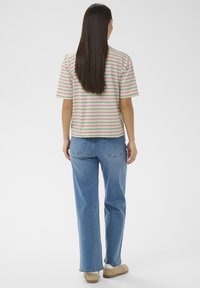 Frau mit langen, glatten Haaren, die ein gestreiftes Kurzarmshirt, blaue Jeans und beige Turnschuhe trägt, steht mit dem Rücken zur Kamera vor weißem Hintergrund.