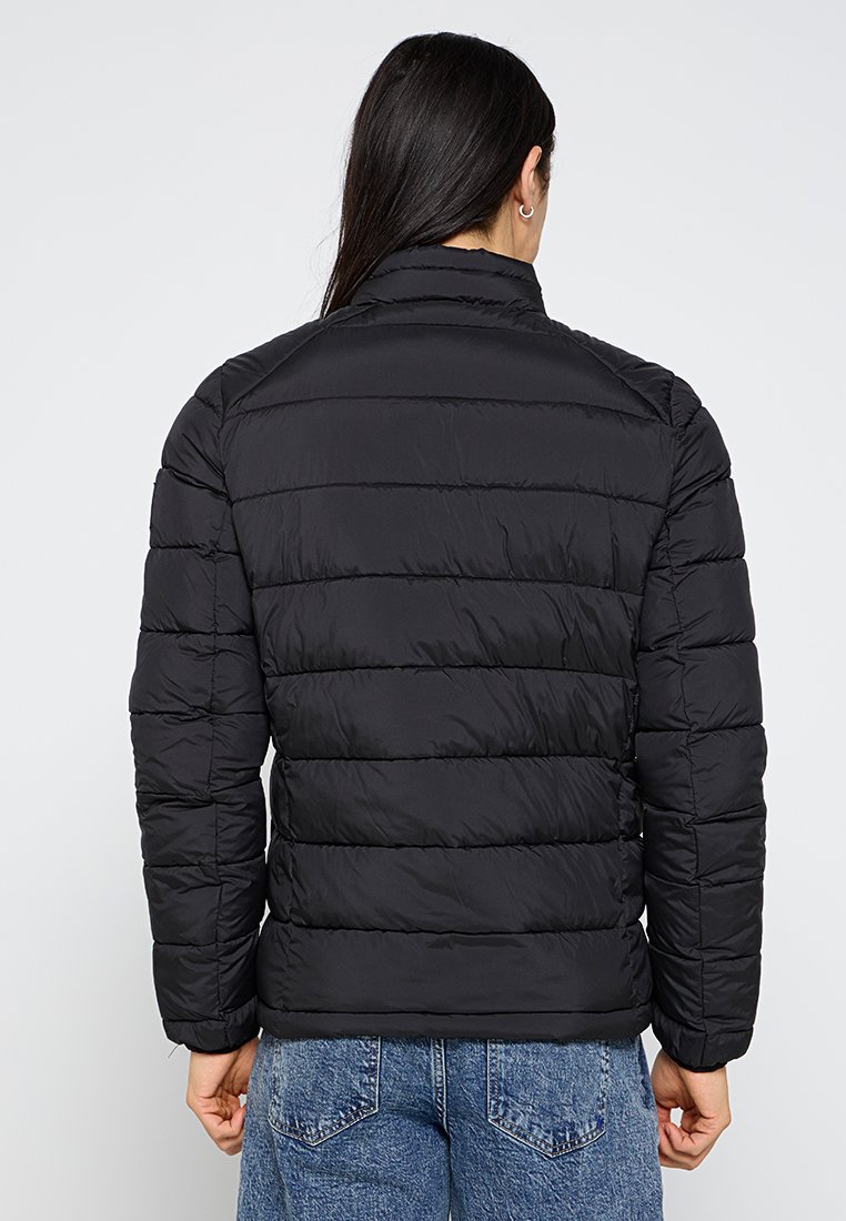 Chaqueta acolchada negra con costuras horizontales, cuello alto y mangas largas. Textura suave, diseño ligero. Lleva puesta sobre unos vaqueros azules.