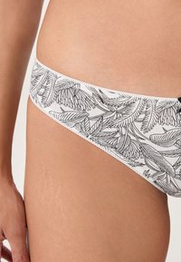 Close-up of a person’s hip wearing white underwear with a black botanical leaf pattern and scalloped edges.