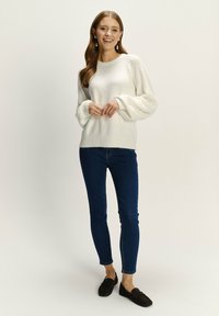 Witte fluffy trui met lange mouwen, blauwe skinny jeans en brown loafers. Neutrale achtergrond benadrukt de texturen en kleuren van de outfit.