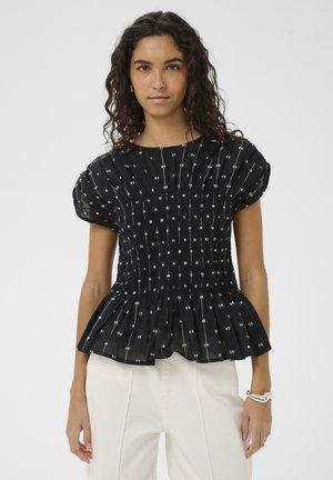 Mujer con cabello rizado, vistiendo una blusa negra de manga corta con fruncidos y lunares blancos, y pantalones blancos de talle alto, posando frente a un fondo liso.