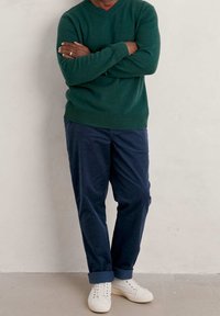 Pull vert en laine tricotée avec poignets et ourlet côtelés, associé à un pantalon chino bleu marine et des baskets blanches, posé devant un fond neutre.