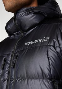 Juodas pūkinis švarkas su blizgiu paviršiumi, turintis gobtuvą, priekinį užtrauktuką ir dvi užtraukiamas kišenes. Ryškiai matomas logotipas "NORRØNA".