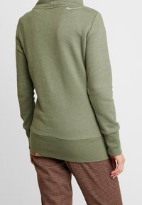 Persona che indossa una felpa verde oliva con testo bianco sul collo e pantaloni a quadri marroni, mostrata di spalle su uno sfondo neutro.