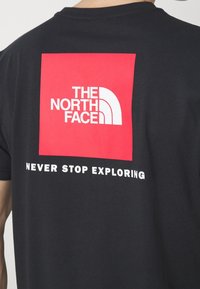Zblízka na zadní část černého trička s červeným čtvercovým logem a bílým textem "THE NORTH FACE" nad sloganem "NEVER STOP EXPLORING".