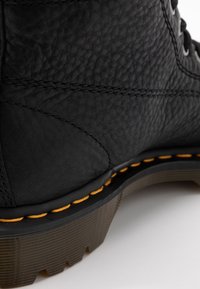 Gros plan d'une botte en cuir noir texturé avec une semelle épaisse crantée et une couture jaune distinctive le long du bord de la semelle.