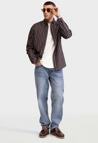 Striped button-up shirt in dark and light tones, paired with a white t-shirt, light blue jeans, and brown lace-up shoes.