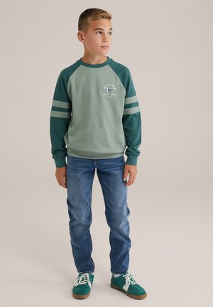 Ragazzo in piedi, che indossa una felpa verde e grigia, jeans blu e sneaker verdi, che guarda a sinistra su uno sfondo semplice.