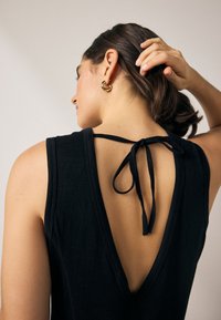 Zwart mouwloos topje met een diepe V-rug en verstelbare striksluiting. Glad stof met zichtbare stikdetails. Gouden oorring als accent.