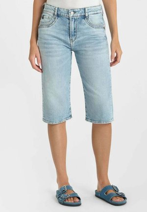 Lichtblauwe denim bermudashorts gedragen door een staande persoon, gecombineerd met blauwe sandalen met gesp en een wit topje.