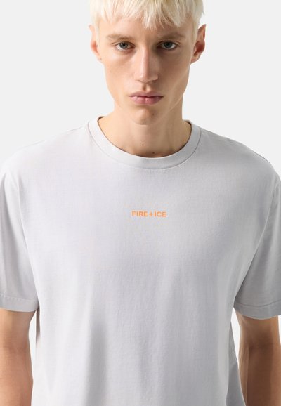 Jeune personne aux cheveux blonds courts portant un T-shirt gris clair uni avec le texte orange « FIRE + ICE » centré sur la poitrine.