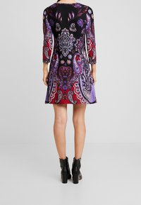 Femme portant une robe mi-longue aux motifs floraux et cachemire violet, rouge et noir, ainsi que des bottines noires, debout de dos.