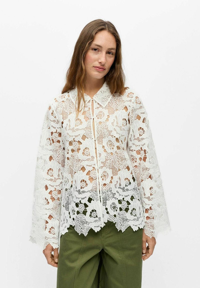 Femme portant un chemisier blanc à manches longues en dentelle florale avec des bords festonnés et un pantalon vert olive sur un fond blanc uni.