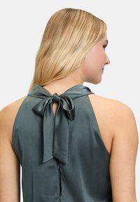 Green satin top featuring a back bow tie accent and a textured surface. The design has a high neckline and a zipper closure.