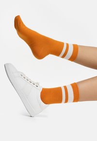 Osoba nosząca pomarańczowe skarpetki z białymi paskami, jedna stopa w białym trampku, druga stopa uniesiona, bez buta, na białym tle.