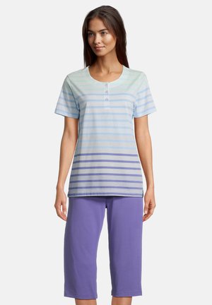 Vrouw draagt een pastel gestreept topje met korte mouwen en knopen en effen paarse capri-broek, staand tegen een effen achtergrond.