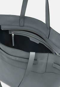 Szara skórzana torba tote o gładkiej fakturze. Posiada zamek błyskawiczny u góry, wewnętrzną kieszeń na zamek i płaskie uchwyty do łatwego przenoszenia. Wnętrze jest ciemne.