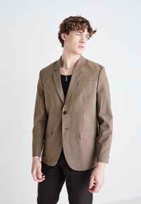 Brauner kariertes Blazer mit einem Knopfverschluss, Reverskragen und zwei aufgesetzten Taschen; getragen über einem schwarzen Hemd mit schlankem Schnitt.