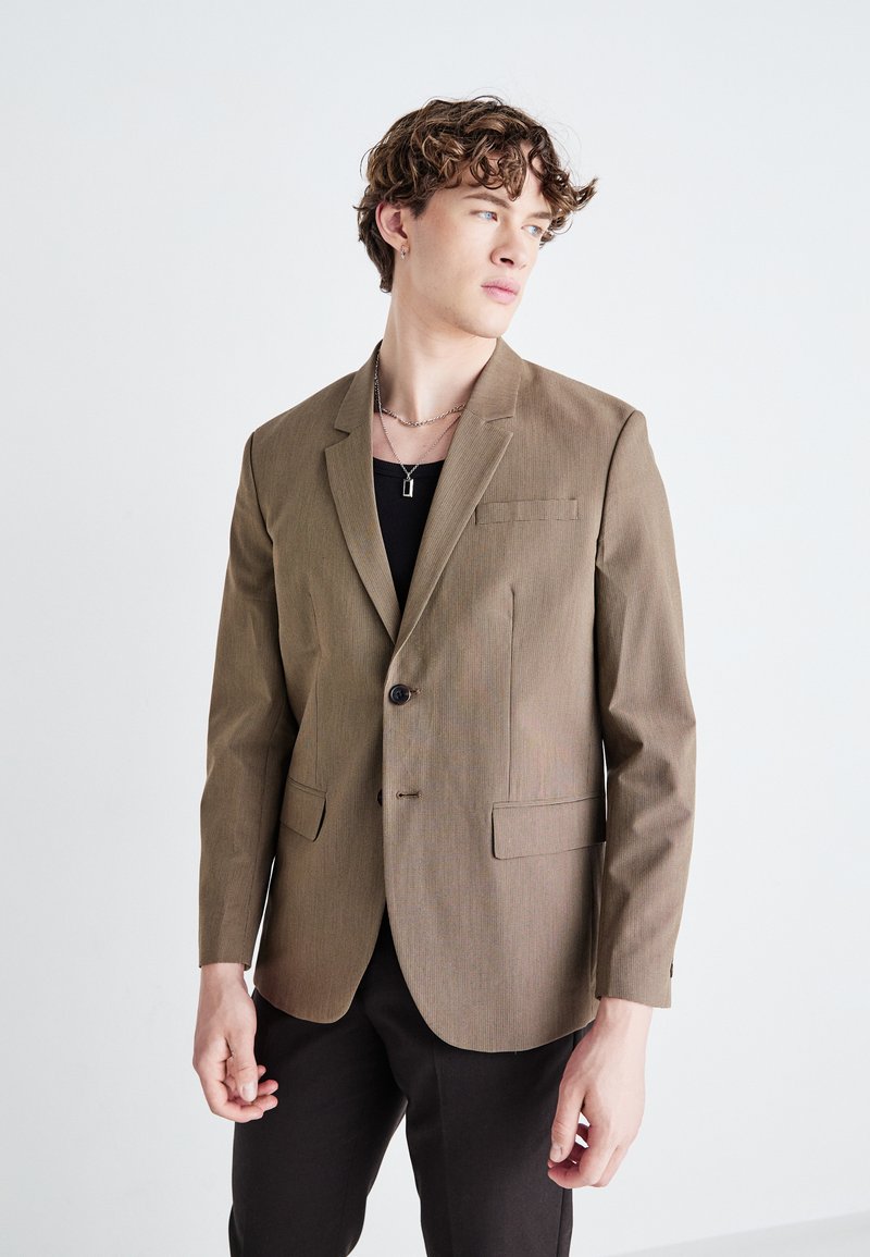 Brauner kariertes Blazer mit einem Knopfverschluss, Reverskragen und zwei aufgesetzten Taschen; getragen über einem schwarzen Hemd mit schlankem Schnitt.