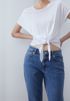 Haut blanc à manches courtes avec un nœud à l'avant, associé à un jean bleu taille haute. Le tissu semble doux et la tenue a une coupe décontractée.