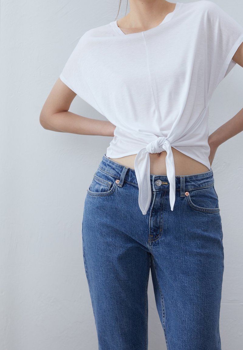 Vit kortärmad topp med en knut framtill, matchad med högmidjade blå jeans. Tyget verkar mjukt, och outfiten har en avslappnad passform.