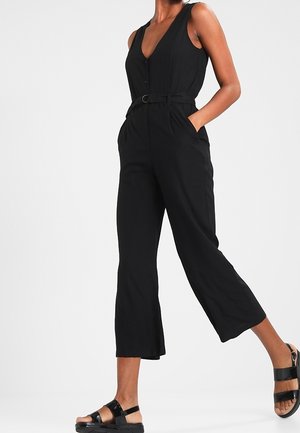 Mono negro sin mangas con escote en v, cintura con cinturón, piernas anchas y recortadas, y bolsillos laterales, hecho de una tela ligera.