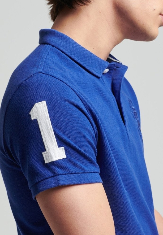 Jeune personne portant un polo bleu avec un écusson blanc numéro un sur la manche, vue de profil sur un fond uni.