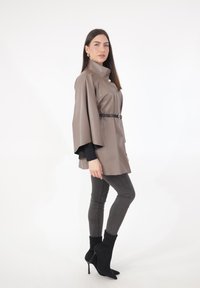 Braunes, kunstledernes Cape-Top mit hohem Kragen, tailliert mit Gürtel, kombiniert mit grauen Skinny-Jeans und schwarzen Stiefeletten mit Absatz.