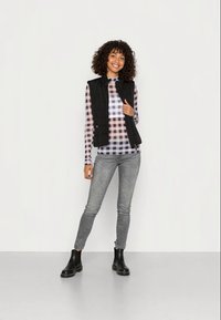 Colete acolchoado preto sobre uma camisa de manga longa xadrez em roxo e branco, combinado com jeans cinza e botas de tornozelo, contra um fundo liso.