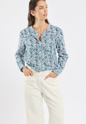 Femme aux longs cheveux bruns portant un chemisier à manches longues bleu et blanc à motif de feuilles et un pantalon taille haute ivoire, les mains dans les poches.