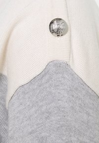 Stickad tröja med en krämfärgad övre del och grå nedre del. En silverknapp med emblemdetalj på axeln ger en dekorativ touch.