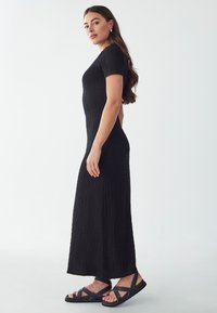 Schwarzes, strukturiertes Maxikleid mit kurzen Ärmeln, figurbetontem Schnitt und seitlichem Schlitz, kombiniert mit schwarzen Sandalen mit Riemen. Einfaches und elegantes Design.
