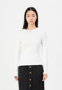 Vit långärmad topp med ribbad textur och scalloped nederkant, som har en liten röd blommaemblem nära halsringningen. Matchas med svart kjol.