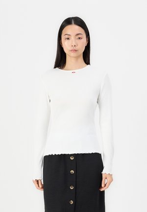 Haut à manches longues blanc avec une texture côtelée et un ourlet festonné, présentant un petit emblème de fleur rouge près du col. Associé à une jupe noire.
