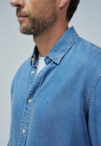 Camisa de mezclilla azul claro con cuello abotonado, dos bolsillos frontales y un pequeño logo bordado en el pecho. Textura suave y diseño informal.