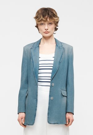 Nő rövid, hullámos hajjal, kopott kék farmerblézert visel fehér és tengerészkék csíkos felső és fehér szoknya fölött.