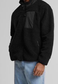 Czarna polarowa kurtka z zamkiem z przodu, wysokim kołnierzem i gładką czarną kieszenią. Luźny krój z teksturowanym, przytulnym wykończeniem.
