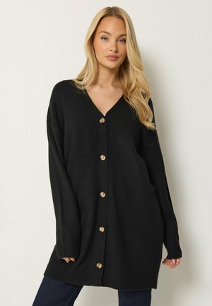 Femme blonde portant un long cardigan noir boutonné et un pantalon foncé, debout devant un fond clair uni.