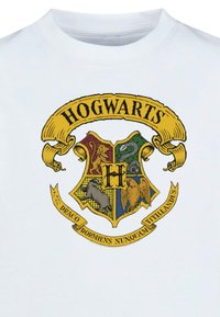 Hvid T-shirt med Hogwarts-våbnet i levende farver. Våbnet inkluderer en løve, en slange, et bæver og en ørn, med gyldne accenter og tekst.