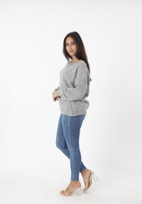 Grijze gebreide trui met gevallen schouders, gecombineerd met blauwe skinny jeans en doorzichtige hakken, gepresenteerd tegen een witte achtergrond.