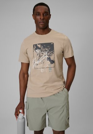 Mężczyzna ma na sobie beżowy t-shirt z wzorem i jasnooliwkowe szorty typu cargo, trzymając w prawej ręce jasnoszarą wielokrotnego użytku butelkę na wodę.