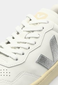 Sneaker bianco in pelle con accenti argentati testurizzati, lacci piatti, dettagli perforati e suola in gomma beige. Elementi di design geometrici visibili.