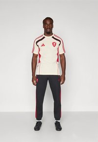 Heren sportoutfit met een crèmekleurige korte mouwen top met rode accenten en logo, gecombineerd met zwarte broek met rode strepen.