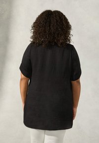 Camicia in lino nera con maniche corte arrotolate, dotata di una superficie testurizzata e di un orlo curvo, vista da dietro.