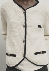 Crèmewitte textuur cardigan met zwarte bies, gouden knopen langs de voorkant en op drie zakflappen, gedragen over een witte top.