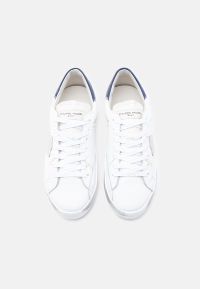 Λευκά δερμάτινα αθλητικά παπούτσια με ομαλή υφή, που διαθέτουν ναυτικό μπλε λεπτομέρεια στη φτέρνα και απλό σχεδιασμό με κορδόνια στο μπροστινό μέρος.