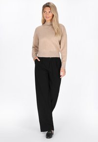 Beige gestrickter Pullover mit hohem Kragen, kombiniert mit schwarzen weit geschnittenen Hosen. Glatte Textur und ribs im Saum und an den Ärmeln.