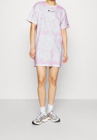 Jasnoróżowa sukienka t-shirt w stylu tie-dye z krótkimi rękawami, z małym czarnym logo na piersi. Połączona z srebrnymi sneakersami i białymi skarpetkami.