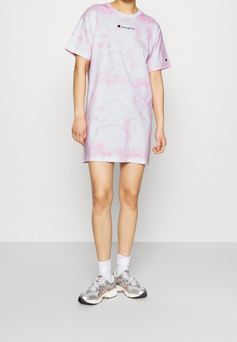 Jasnoróżowa sukienka t-shirt w stylu tie-dye z krótkimi rękawami, z małym czarnym logo na piersi. Połączona z srebrnymi sneakersami i białymi skarpetkami.