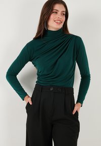 Donker groene top met lange mouwen, een hoge halslijn en een gedrapeerd ontwerp aan de voorkant, gecombineerd met zwarte, hoogopgestelde broek met twee knopen.
